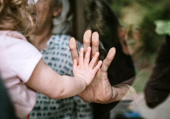 A mother stands with her daughter, visiting senior parents but observing social distancing with a glass door between them.  The granddaughter puts her hand up to the glass, the grandfather and grandmother doing the same.  A small connection in a time of separation during the Covid-19 pandemic.